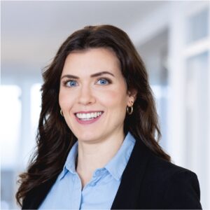 Frau mit langen, dunklen Haaren und blauen Augen lächelt in die Kamera, trägt ein hellblaues Hemd und einen schwarzen Blazer.